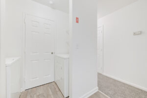 Interior unit linen closet, washer and dryer, racks above dryer, wood-like floors, neutral toned carpeting in hallway.