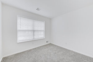 Interior Unit Bedroom, large window, white walls, neutral toned carpeting.